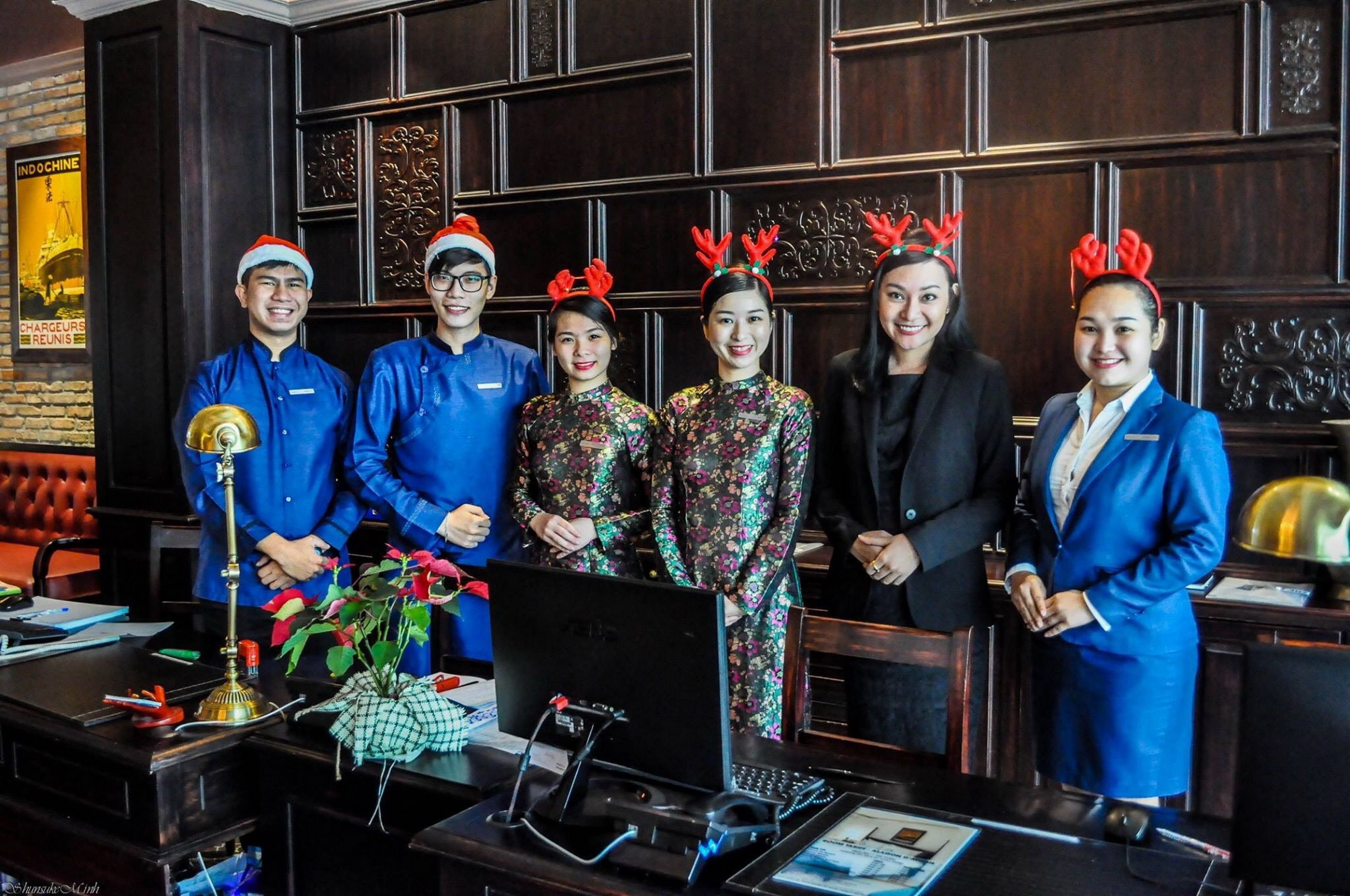 A group of people standing in a hotel lobby with a reception desk, a laptop, and a decorative wall.