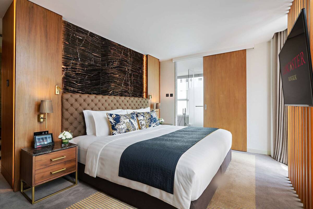 Een moderne hotelkamer met een groot bed, een houten hoofdbord, een nachtkastje met lamp en een televisie aan de muur.