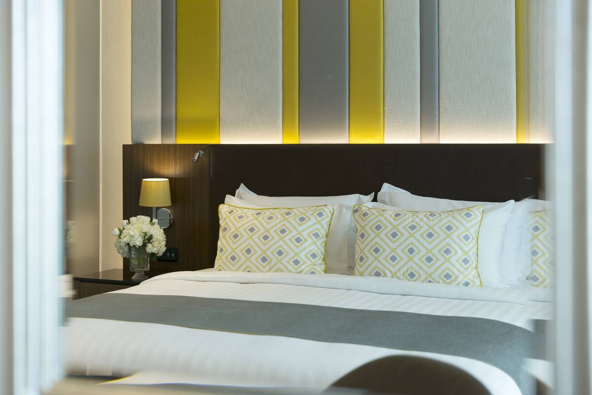 Een opgemaakt bed met gebloemde kussens, een bedlampje en een vaas met bloemen.