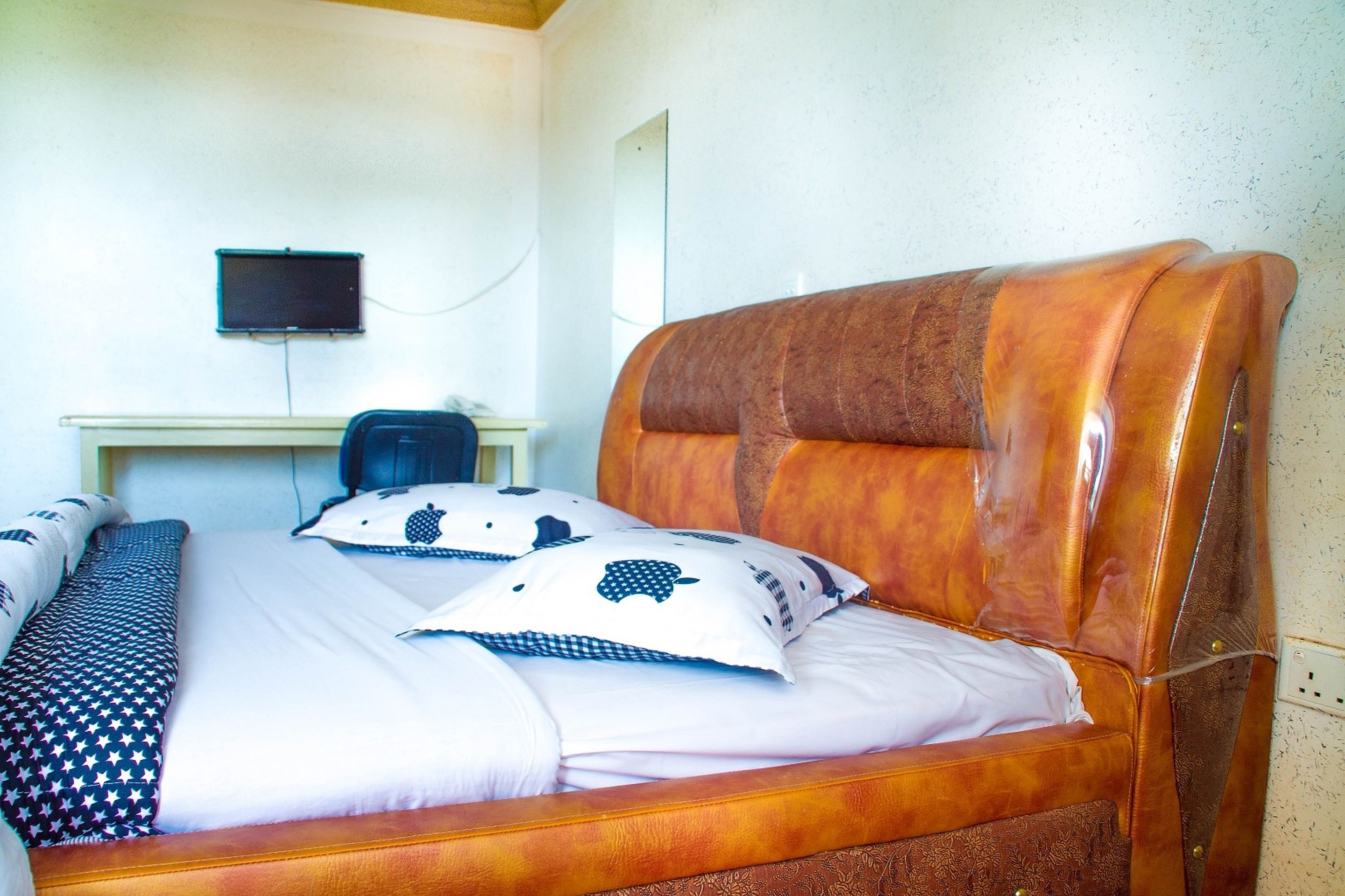 Una cama bien tendida con almohadas estampadas, un cabecero de madera, un escritorio con computadora y una televisión instalada en la pared.