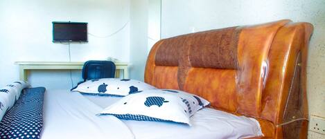 Una cama bien tendida con almohadas estampadas, un cabecero de madera, un escritorio con computadora y una televisión instalada en la pared.