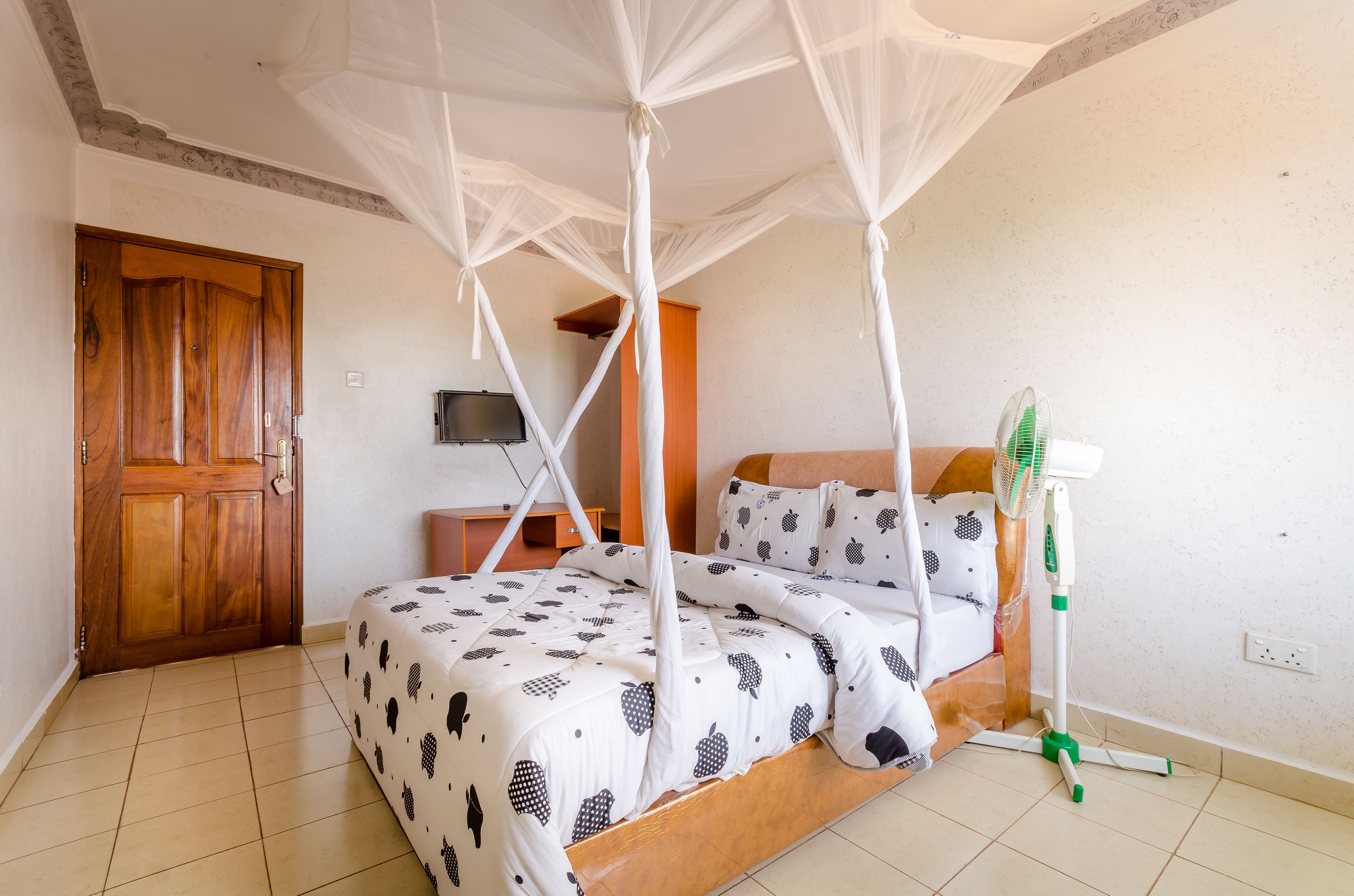A bedroom with a bed, a wooden door, a television, and a standing fan.
