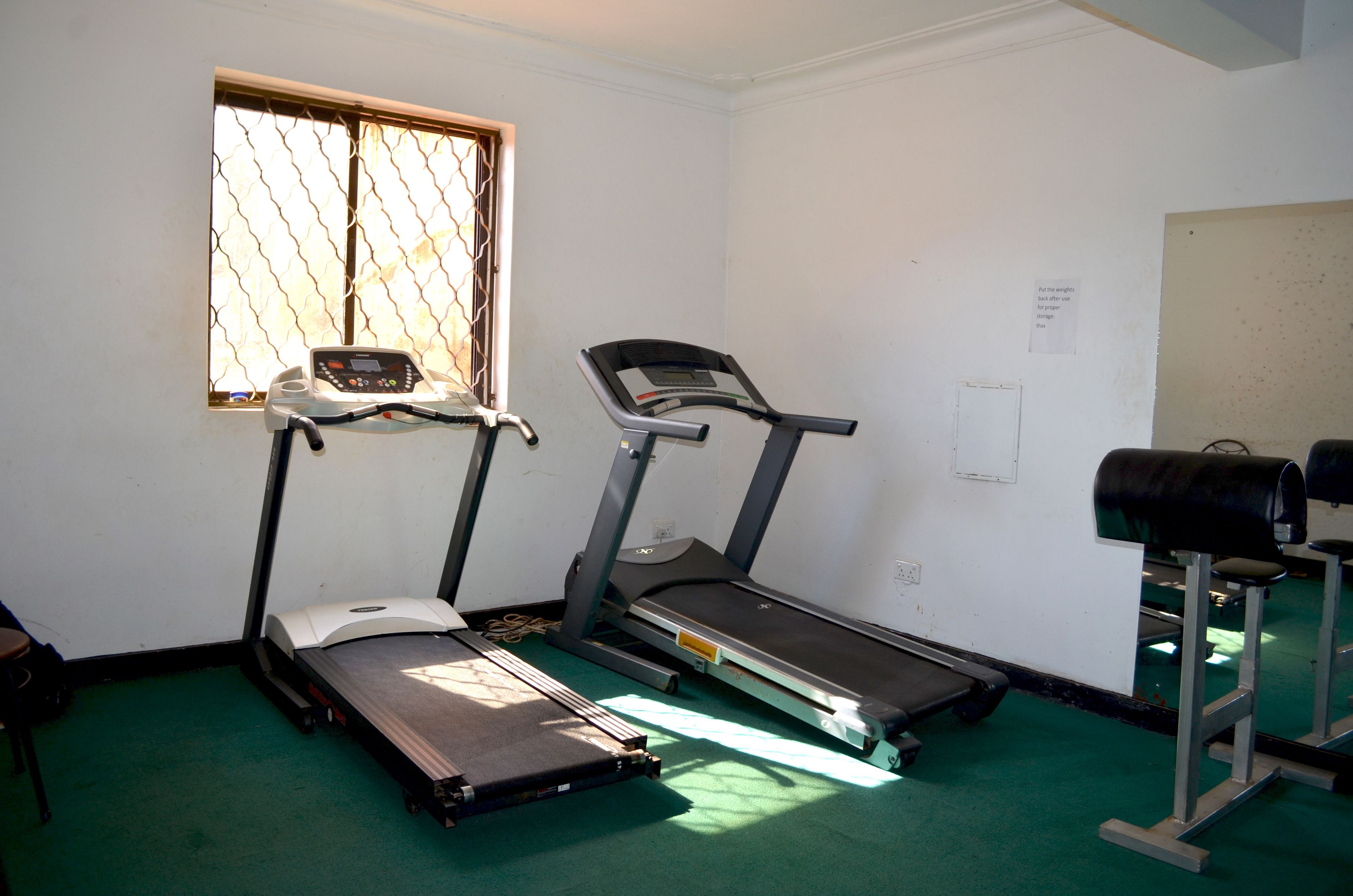 A room with two treadmills, a window with metal bars, and a green carpet.