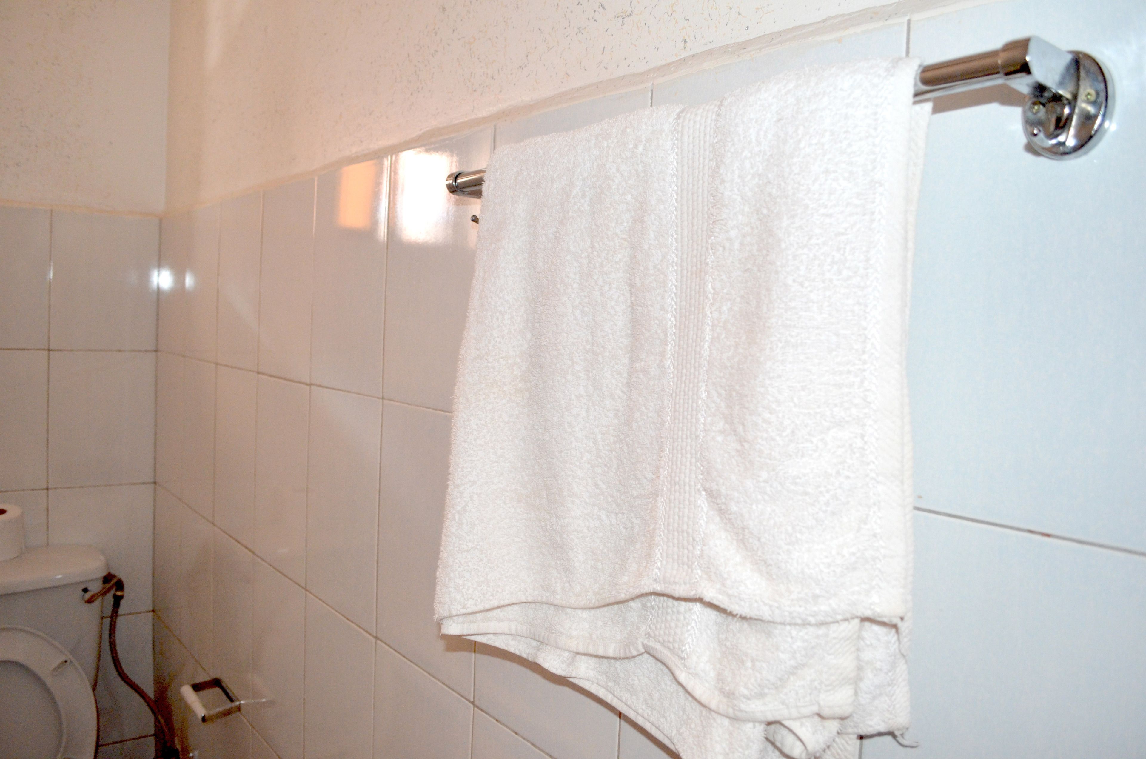 A bathroom with white tiled walls, a white towel hanging on a metal towel rack, and a toilet with a visible pipe.