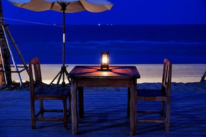 A wooden table with a lantern on it, two chairs, and an umbrella on a wooden deck by the beach at night. - Keeree Waree Seaside Villa (Bang Saphan)