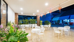 Un patio al aire libre con mesas y sillas blancas, rodeado de plantas en macetas y faroles decorativos.