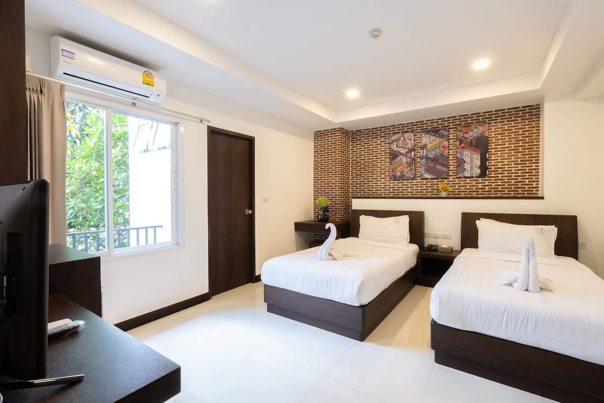 A hotel room with two beds, a TV, a brick accent wall, and a window with a view of greenery.