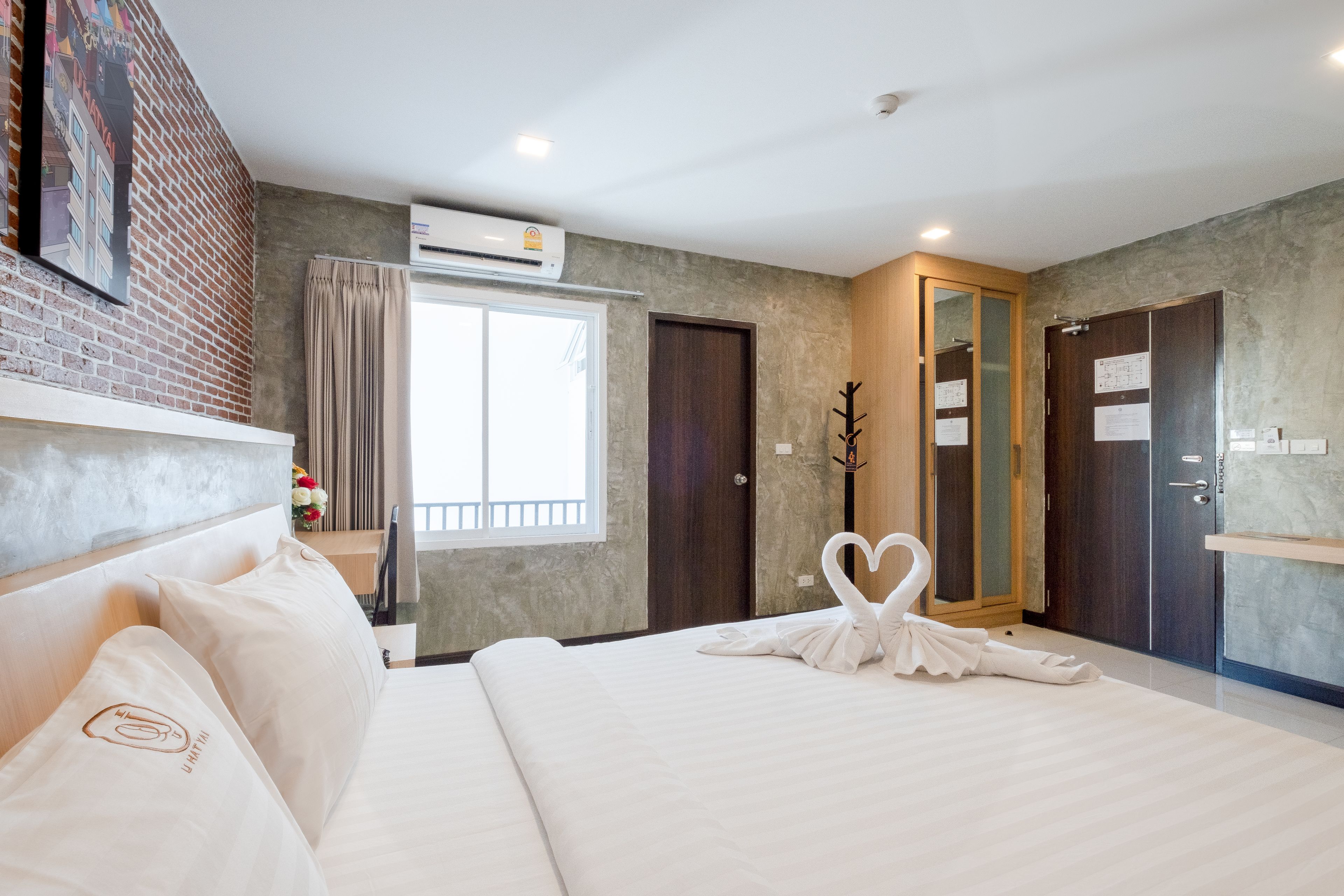A hotel room with a large bed, a wooden headboard, a brick accent wall, a window with curtains, a door with a mirror, and a wall-mounted air conditioner.
