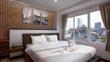 A hotel room with a brick accent wall, a large bed with white bedding, a city view through the window, and a nightstand with a phone and a lamp.