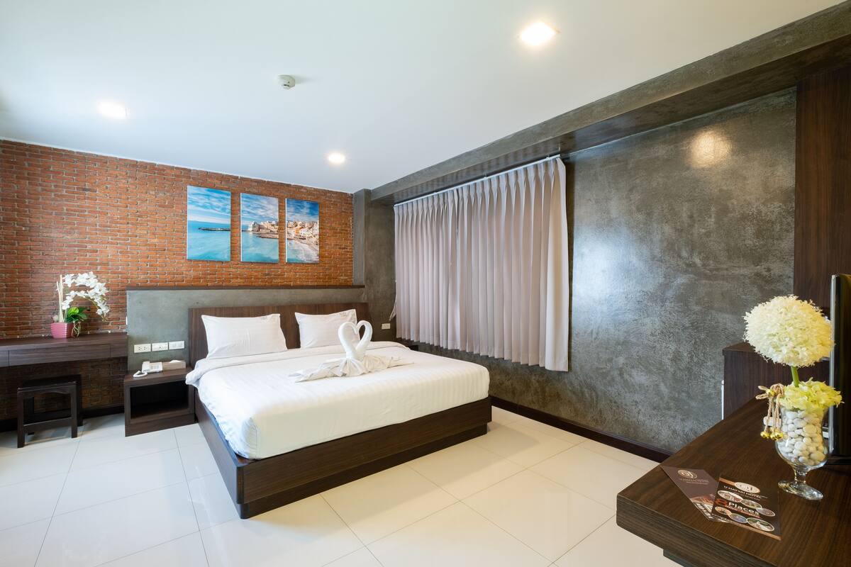 A modern hotel room with a brick accent wall, a large bed, a desk, and a view of the sea through the window.