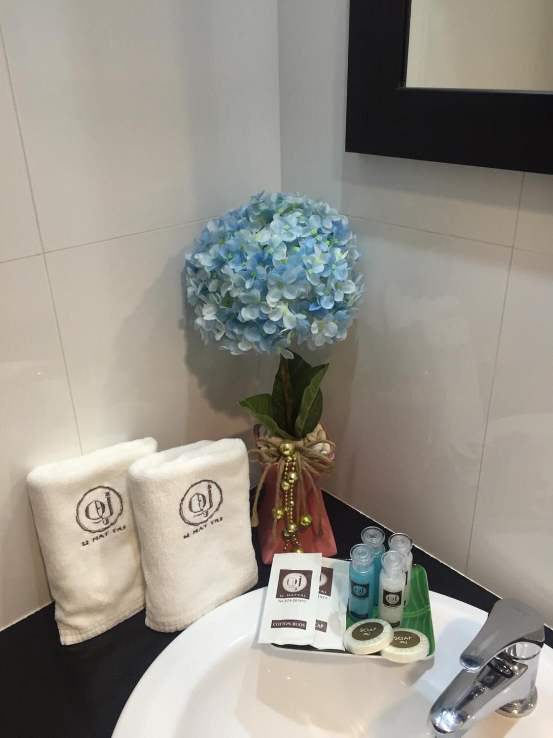 Une salle de bain avec un lavabo blanc, deux serviettes blanches ornées d’un logo, une composition florale artificielle bleue et des articles de toilette, notamment du savon et de la lotion.