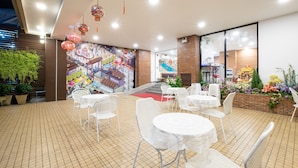 Un área de comedor exterior moderna con sillas y mesas blancas, un mural de una calle comercial y faroles rojos colgados del techo.