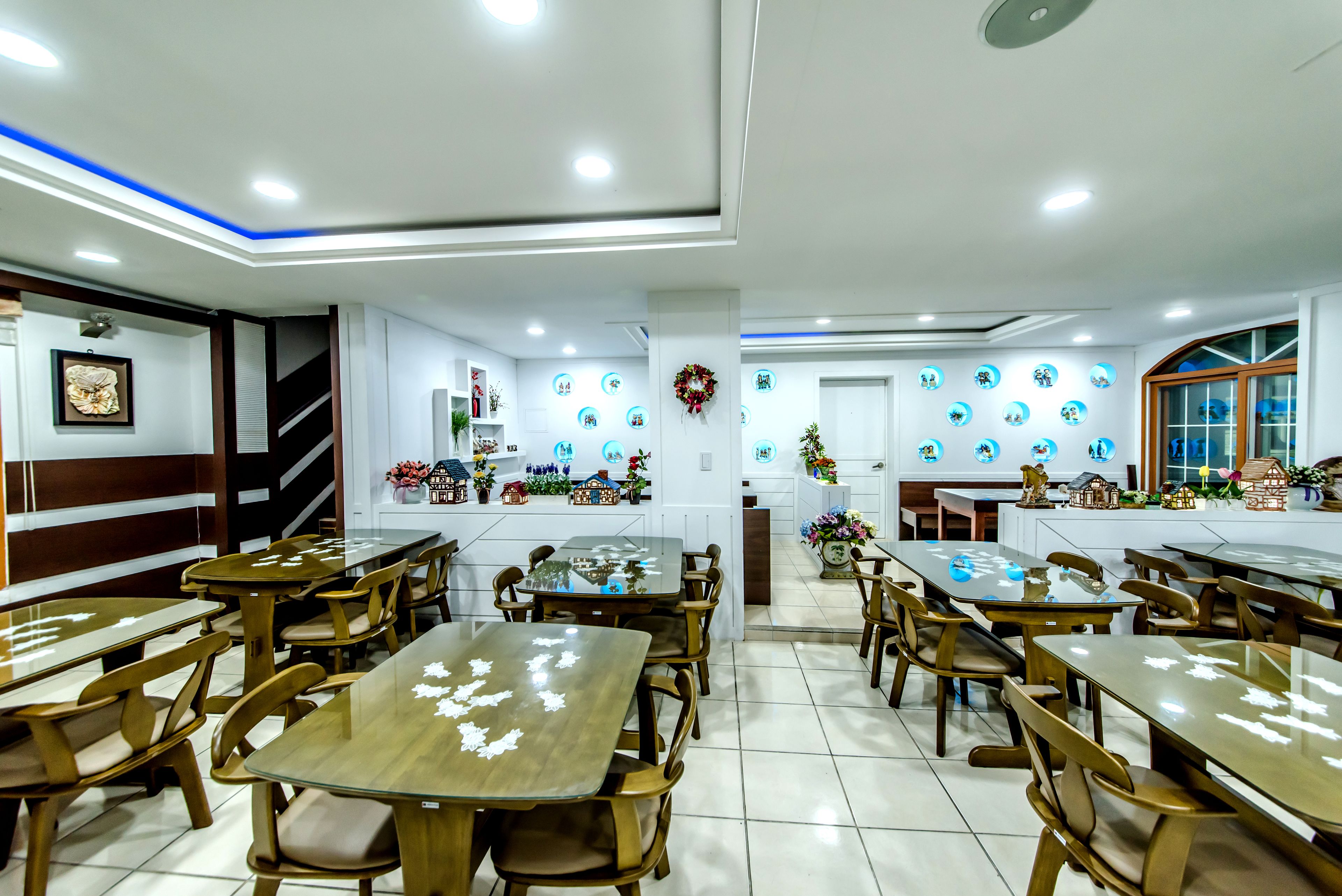 A restaurant with multiple wooden tables and chairs, a white tiled floor, and a wall decorated with circular plates and a wreath.