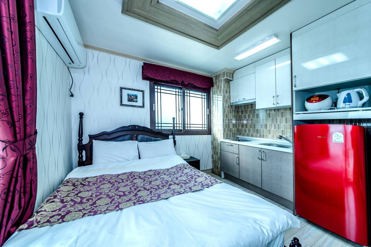 Une chambre avec un lit, une table de chevet, une fenêtre avec des barreaux, un réfrigérateur rouge et un coin cuisine avec des placards et un évier.