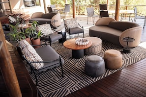A covered outdoor seating area with sofas, chairs, and tables, surrounded by wooden flooring and potted plants. - Bona Bona Game Lodge (Maquassi Hills)
