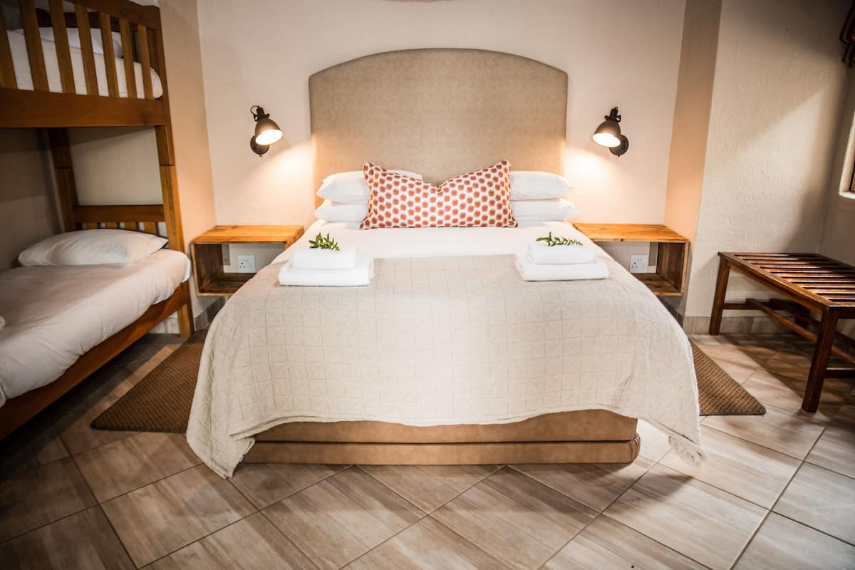 Un lit double avec une tête de lit, deux tables de chevet avec des lampes, un banc en bois et un lit superposé sur le côté gauche.