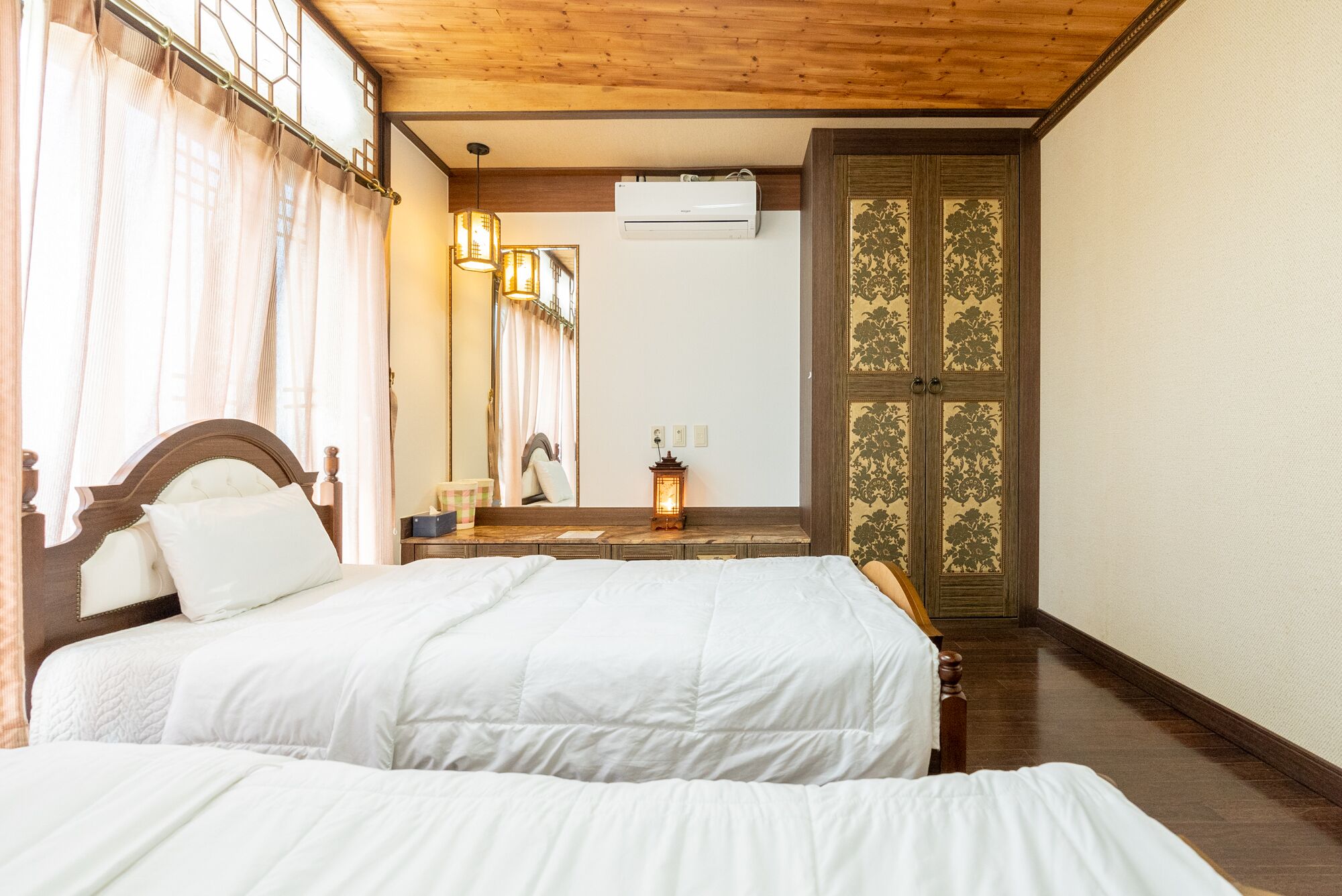 Habitación de hotel con dos camas, techo de madera, un espejo, unidad de aire acondicionado y una puerta tradicional con motivos florales.