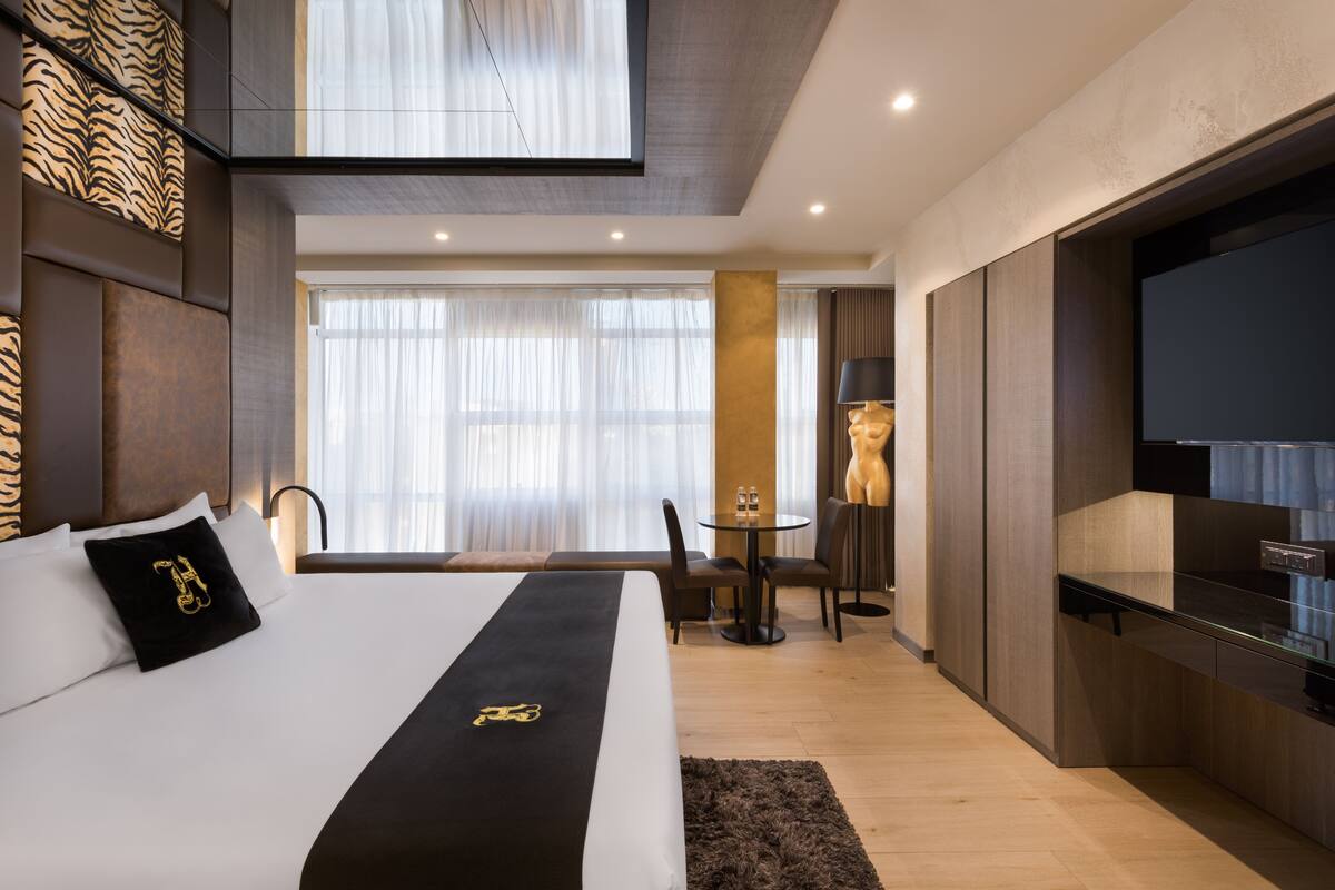 A modern hotel room with a large bed, a flat-screen TV, a dining area, and a view of the city.