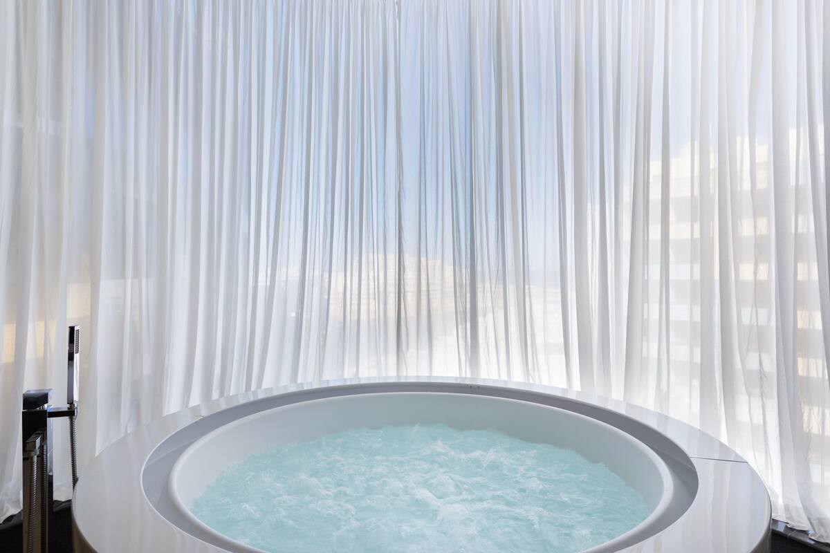 A hot tub with a view of a building through sheer curtains.