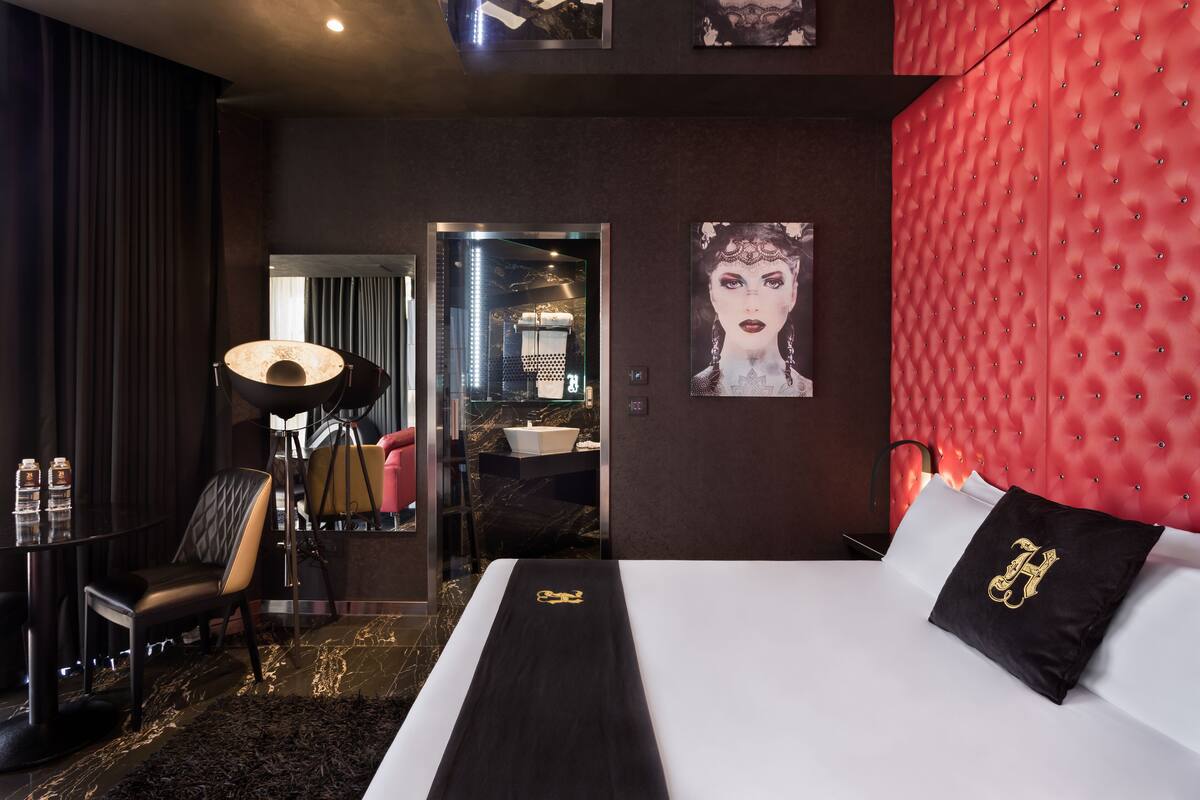 A modern hotel room with a large bed, a red tufted headboard, a black and white striped bed runner, a black pillow with a gold emblem, a black armchair, a floor lamp, and a view of the bathroom through a glass door.