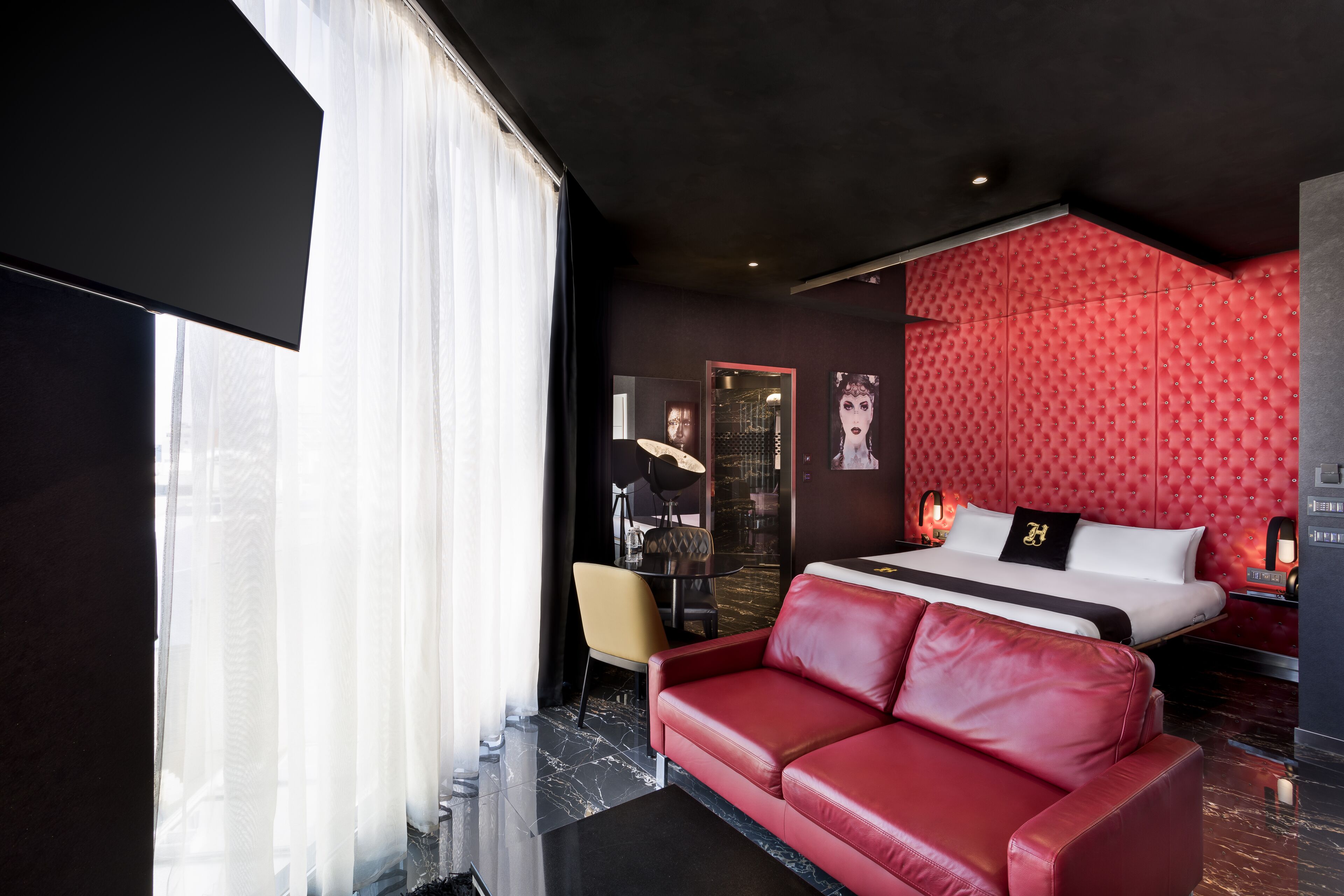 Ein modernes Hotelzimmer mit einem großen Bett, einer roten Ledersofa, einem Esstisch und Blick auf die Stadt durch große Fenster.
