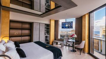 Habitación de hotel moderna con una cama grande, un balcón con vistas a la ciudad, una zona de comedor y una pantalla de televisión mostrando un menú.