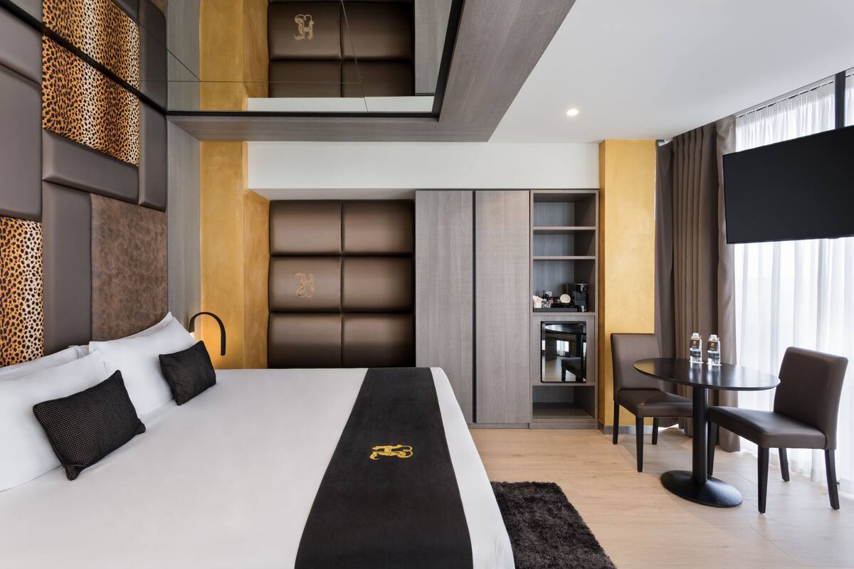 A modern hotel room with a large bed, a flat-screen TV, a dining area, and a wardrobe.