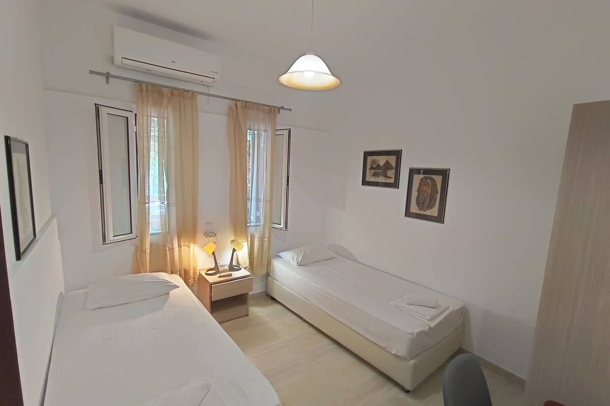 A small, clean room with two single beds, a bedside table with lamps, a chair, and a window with curtains.
