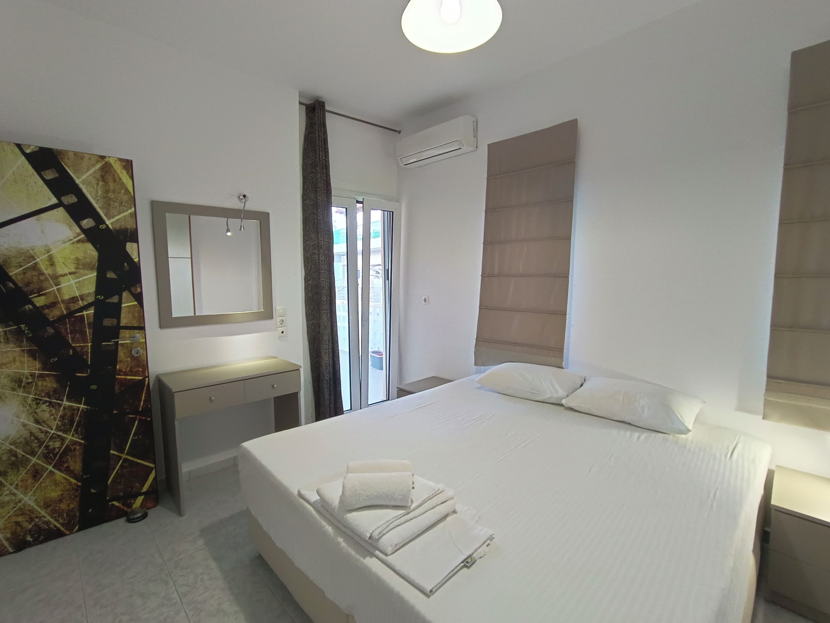 A hotel room with a bed, a bedside table, a mirror, an air conditioning unit, and a view of the outside.