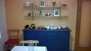 Un comedor con una mesa dispuesta para dos personas, un armario azul con diversos utensilios de cocina, y repisas superiores con fotografías enmarcadas y utensilios de cocina.