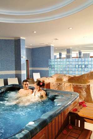A hot tub with a wooden bench and flowers, surrounded by blue patterned walls and a tiled floor. - Alua Helios Bay (Nessebar)