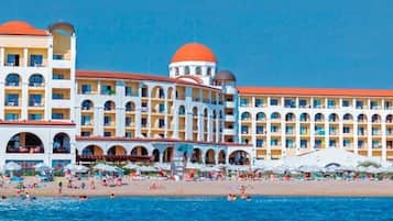 Een groot hotel met meerdere balkons, een strand ervoor en een helderblauwe lucht.