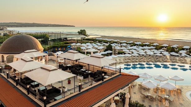 A rooftop bar with outdoor seating overlooking a pool and beach. - Alua Helios Bay (Nessebar)
