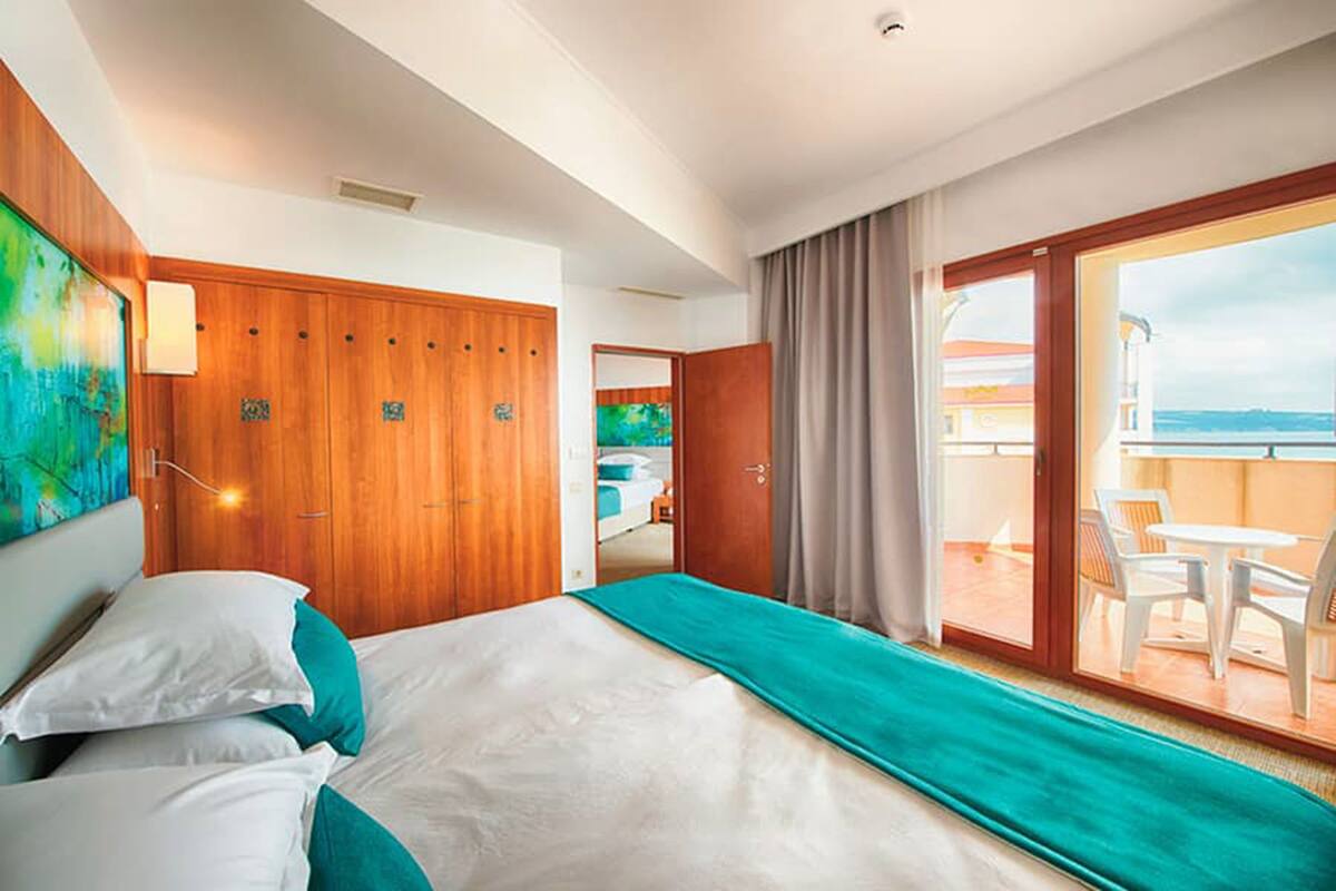 A hotel room with a bed, a wooden wardrobe, a balcony with a table and chairs, and a view of the sea.