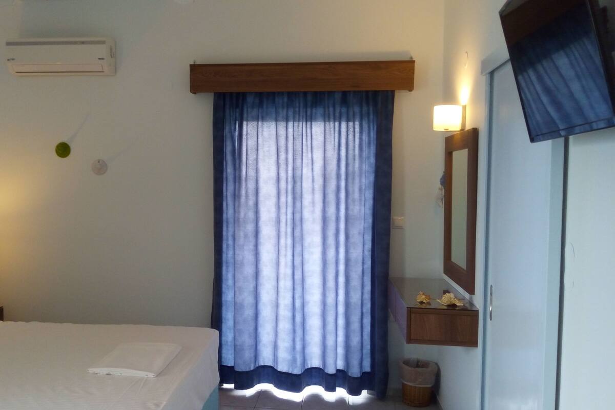 A hotel room with a bed, a nightstand, a wall-mounted air conditioner, a window with blue curtains, a bedside lamp, and a small shelf with decorative items.