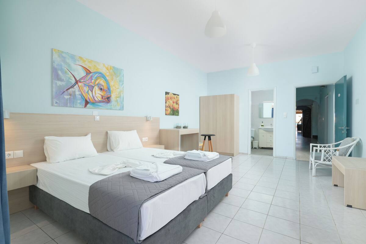 A modern hotel room with a large bed, a desk, and a chair. There is a painting on the wall and a door leading to another room.
