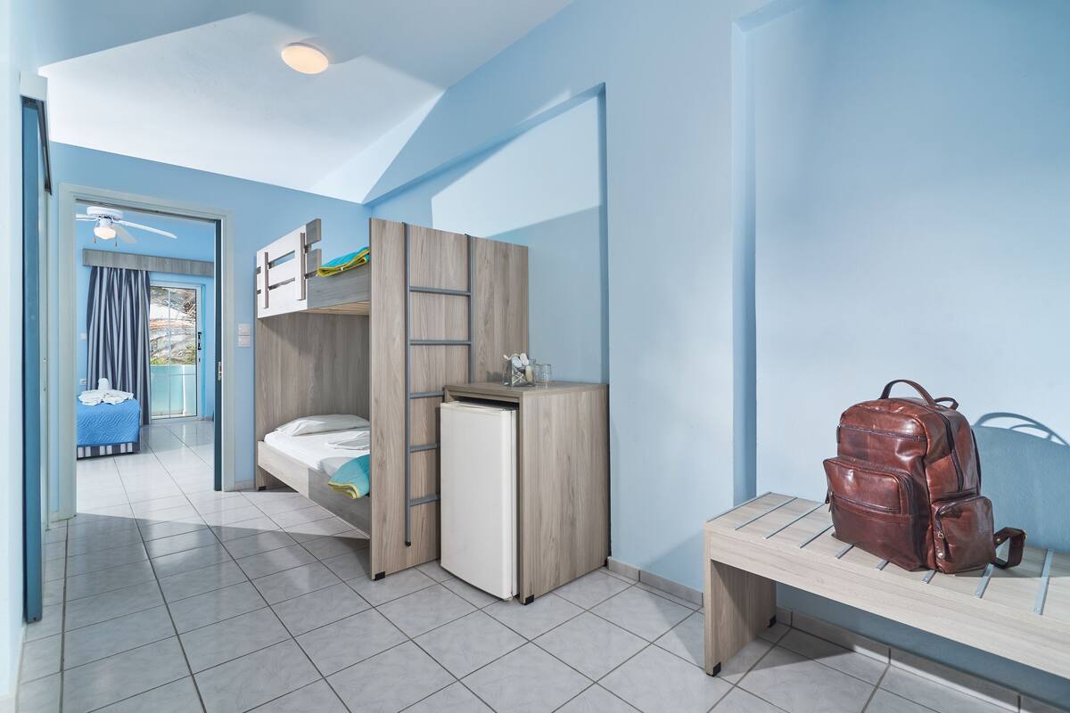 A room with a bunk bed, a small refrigerator, a suitcase on a bench, and a door leading to a balcony.