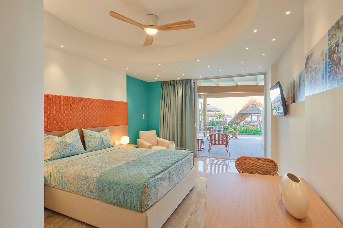 A bedroom with a bed, a ceiling fan, a table with a vase, and a view of the outside.