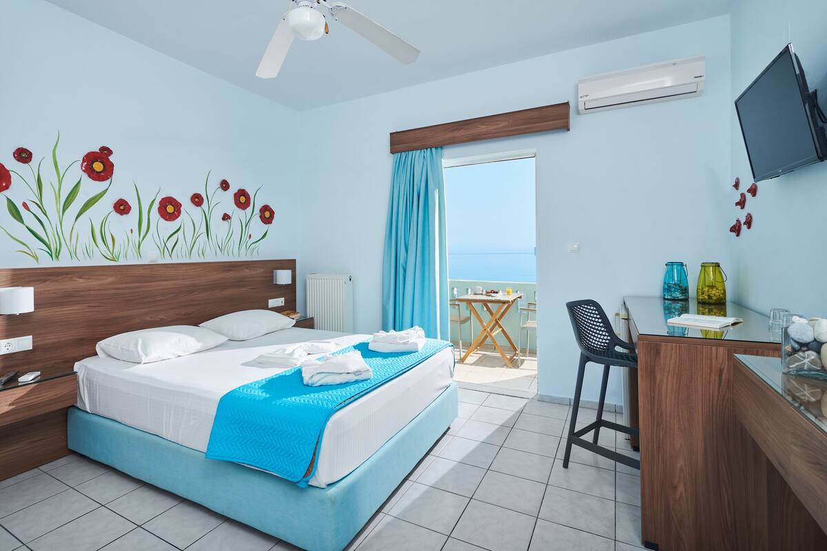 A hotel room with a bed, a desk, a chair, a TV, and a view of the sea.