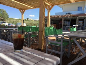 Un espacio al aire libre con mesas y sillas, un vaso de bebida helada, y un edificio con un letrero que dice "APTERA HOTEL".