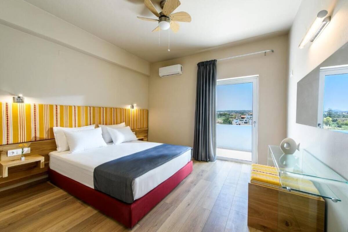A hotel room with a bed, a ceiling fan, a window with curtains, a bedside table, and a view of the outside.