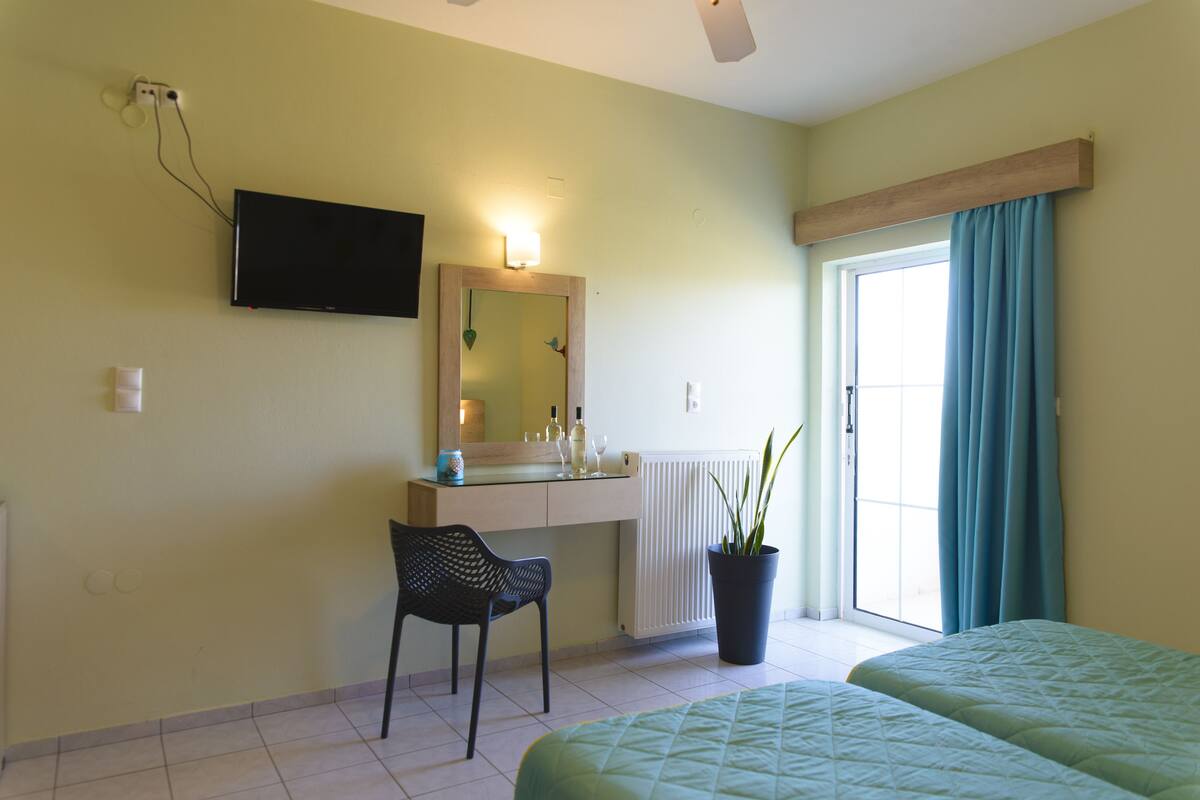 A hotel room with a bed, a chair, a desk with a mirror, a TV mounted on the wall, and a window with curtains.