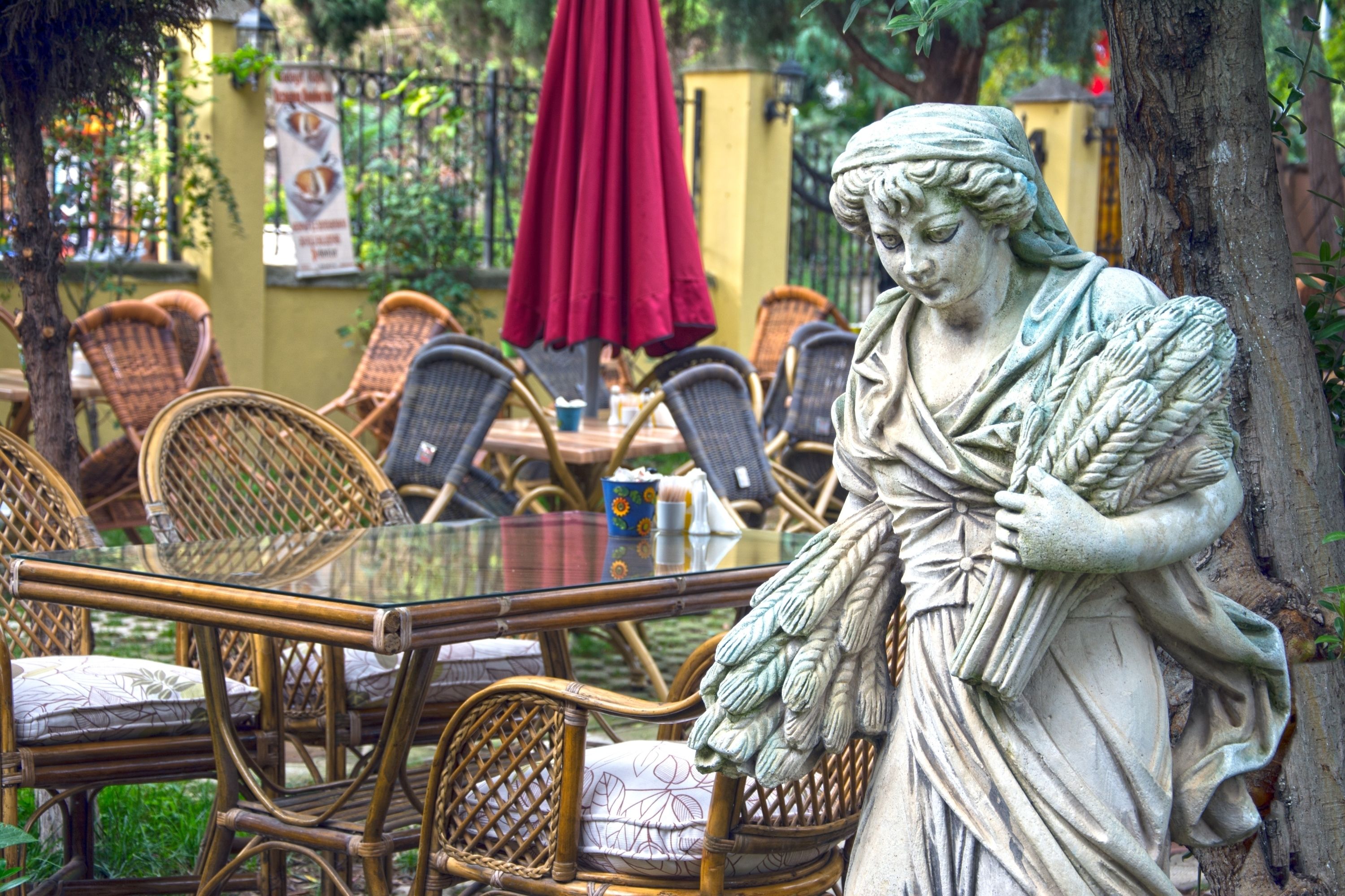 Una estatua de una mujer sosteniendo una rama, rodeada de sillas de mimbre y una mesa de vidrio en un entorno al aire libre.
