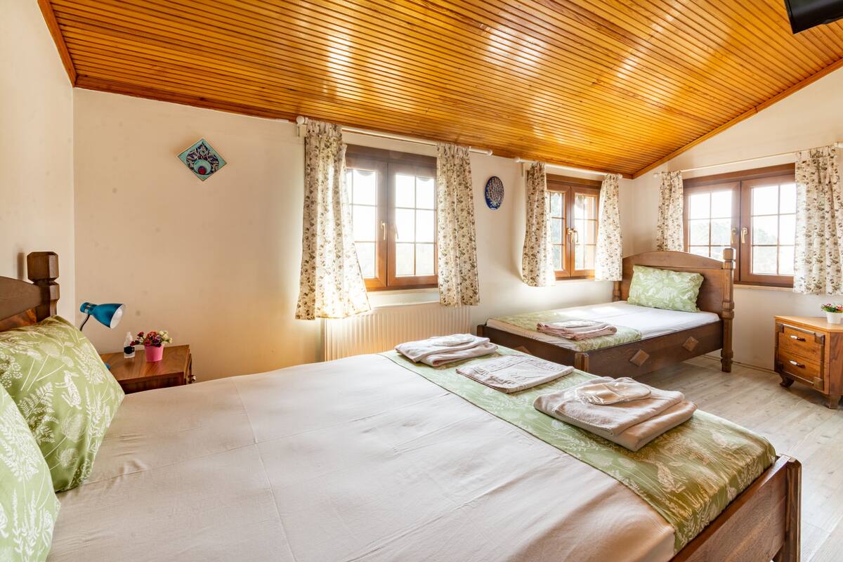 A room with two beds, wooden ceiling, floral curtains, and a small table with a lamp.