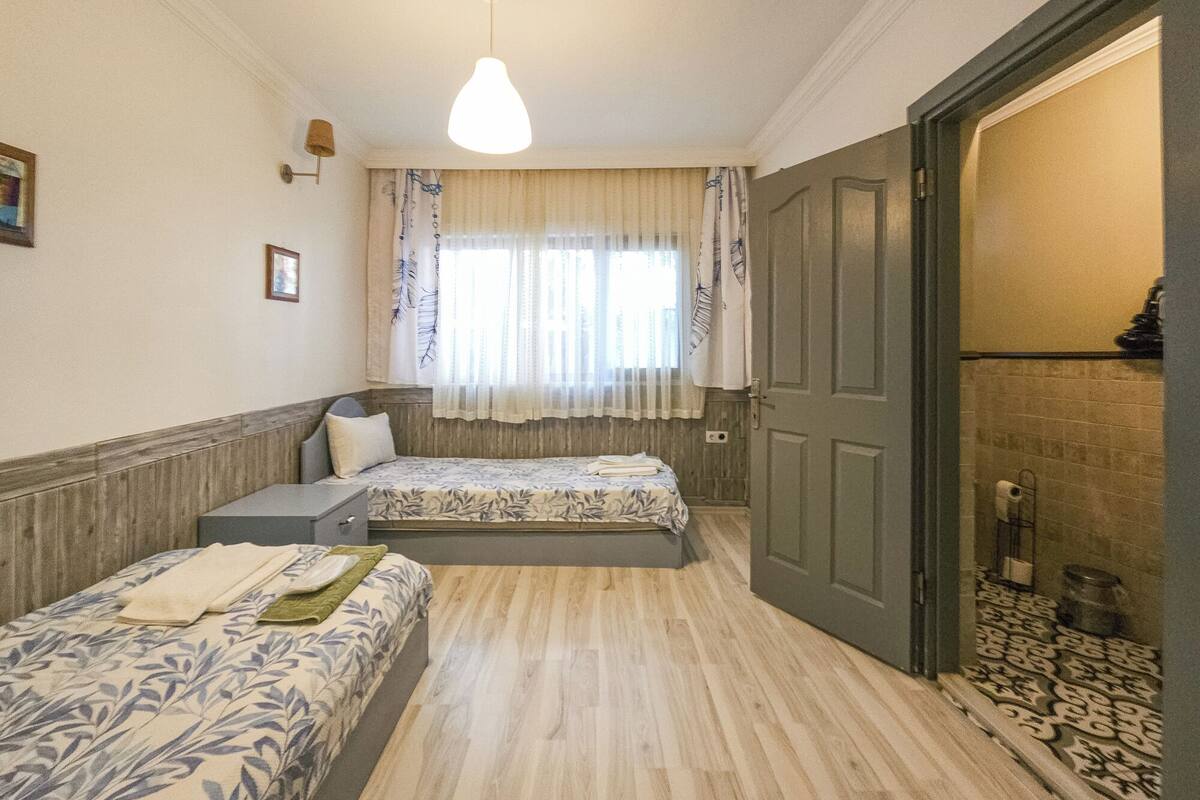 A room with two beds, a wooden floor, a window with curtains, and a door leading to another room.
