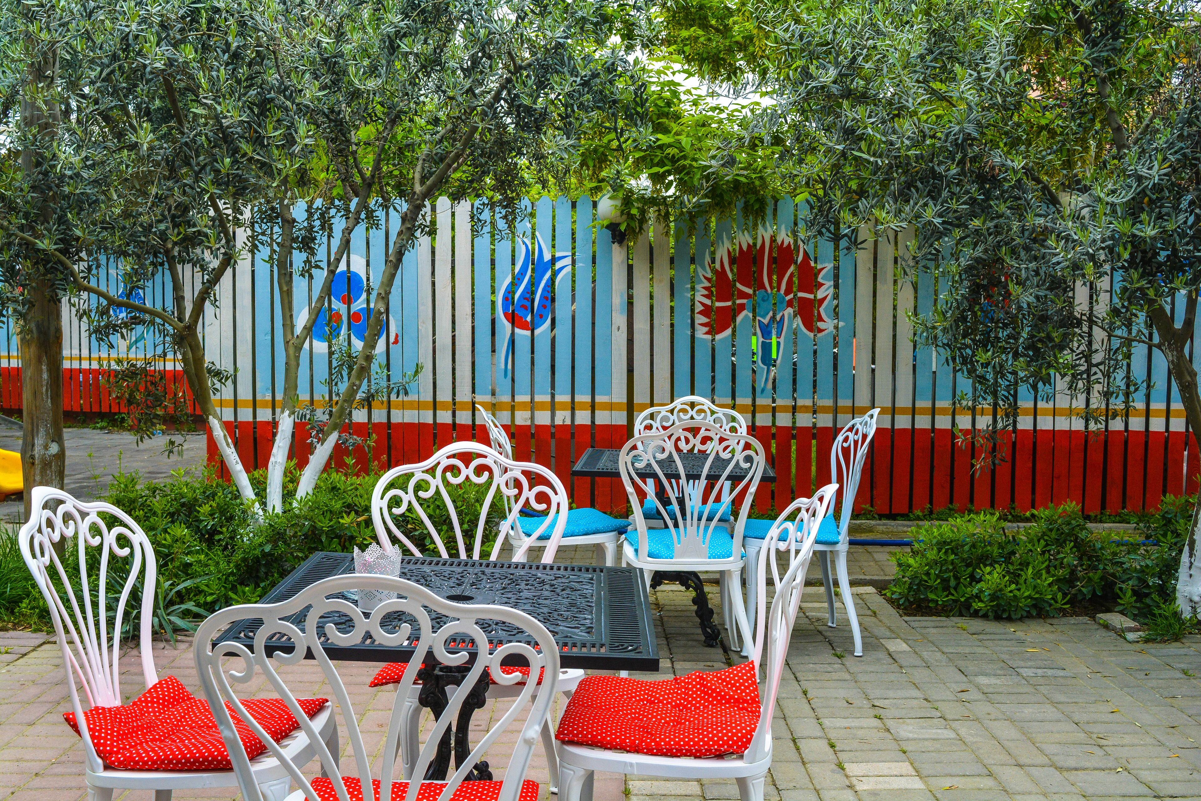 Outdoor seating with white metal chairs and red cushions, a blue table, and a mural with colorful designs on a fence.