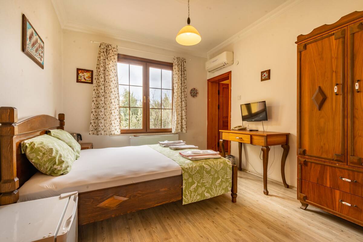 A bedroom with a bed, a wooden wardrobe, a desk with a television, and a window with curtains.
