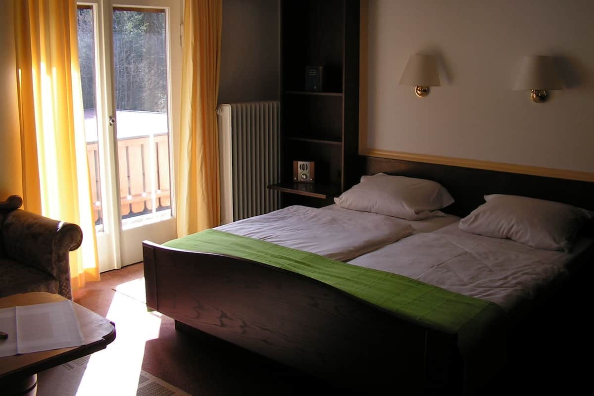 A hotel room with a double bed, a chair, a small table, a radiator, and a view of the outdoors through a sliding door.