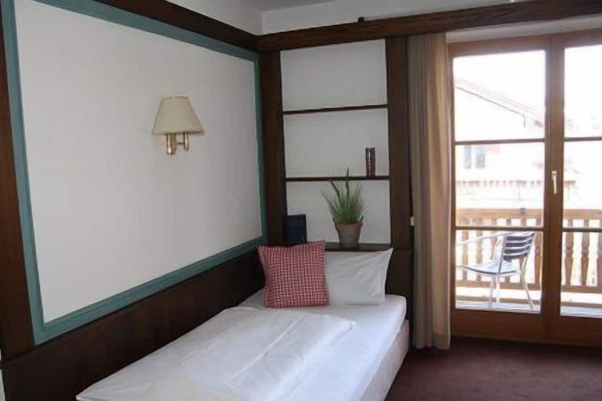 A hotel room with a bed, a nightstand, a lamp, a potted plant, and a balcony with a chair.