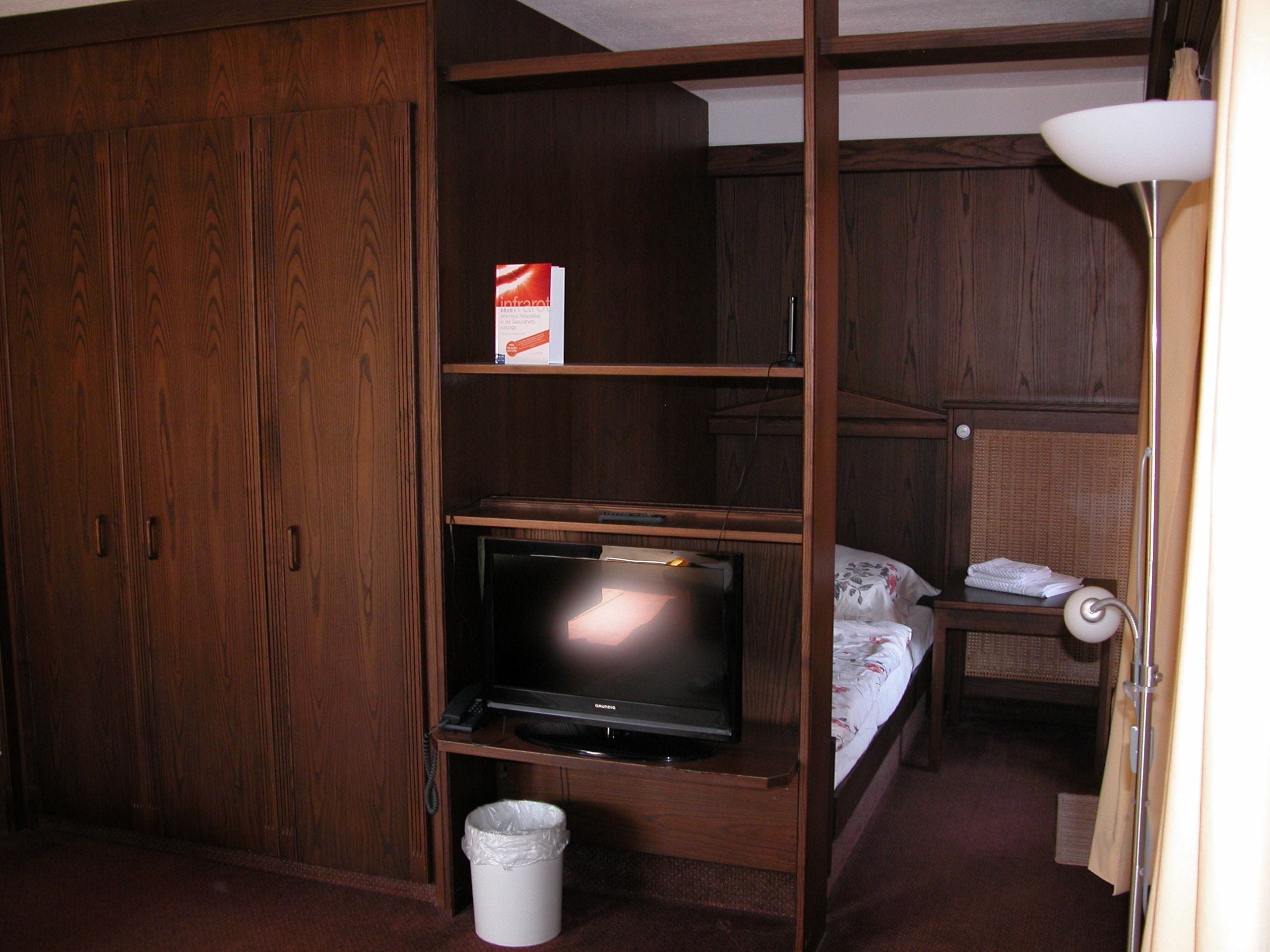 A compact room with a wooden wardrobe, a television on a wooden stand, a small bedside table with a lamp, and a bed with a floral bedspread.